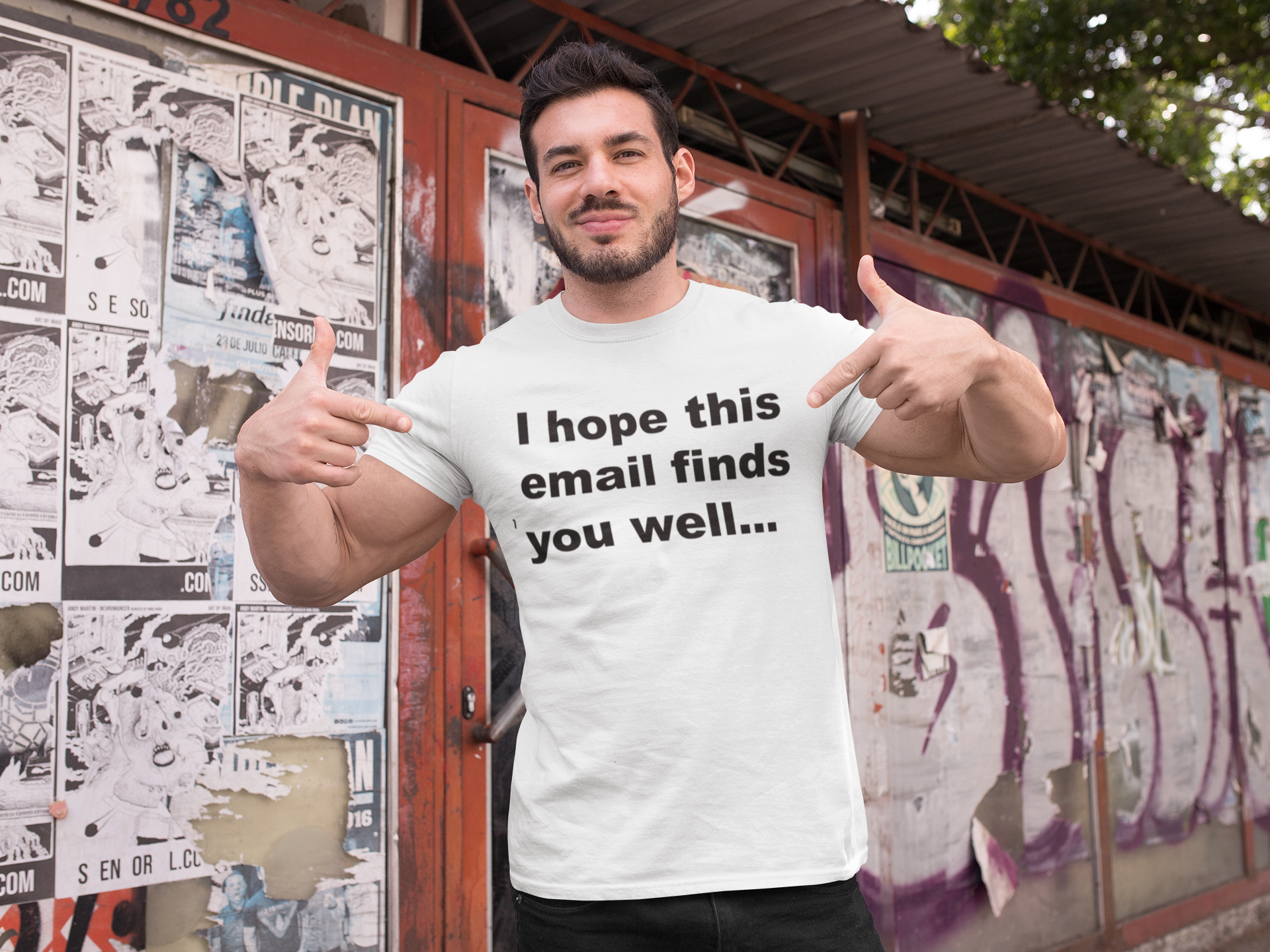 I Hope This Email Finds You Well 100% Organic Cotton T Shirt On Man wearing a white t-shirt with text, giving a thumbs-up in front of a graffiti-covered wall.