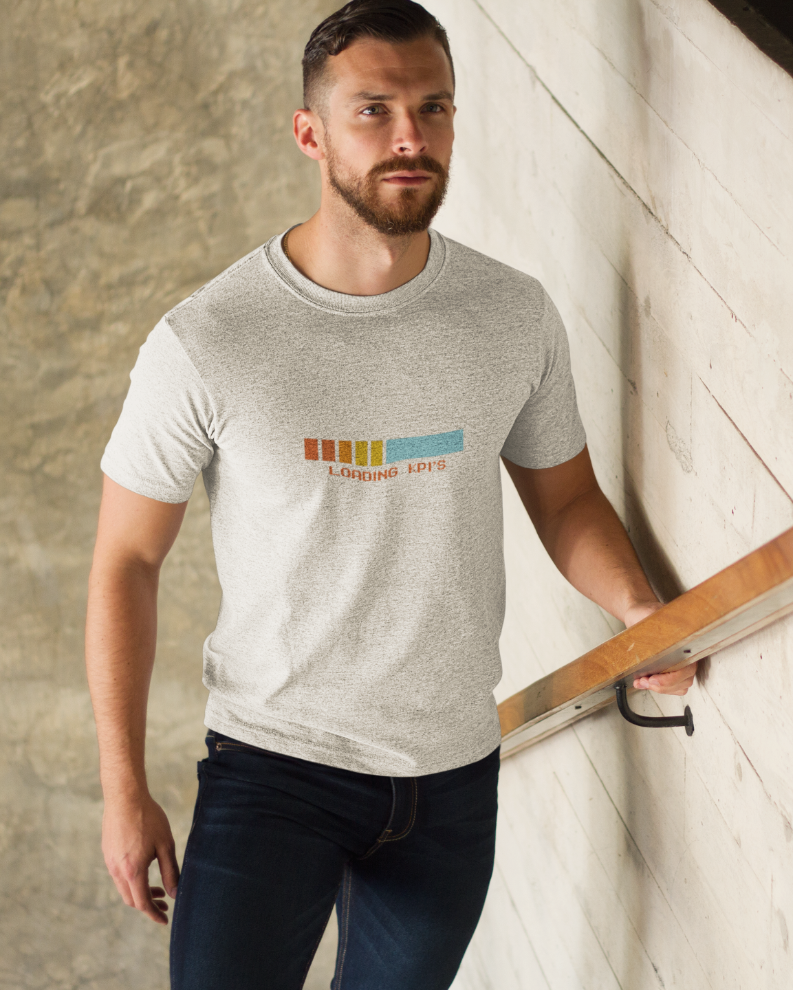 Loading KPI's Humourous T-Shirt Man wearing a gray t-shirt with colorful text design, standing indoors against a textured wall.