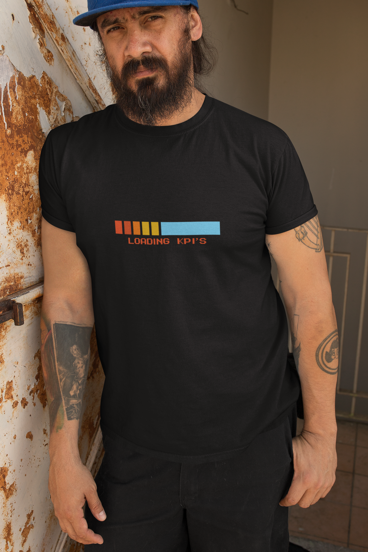 Loading KPI's Humourous T-Shirt Man wearing a black t-shirt with colorful text design, leaning against a rusty wall.