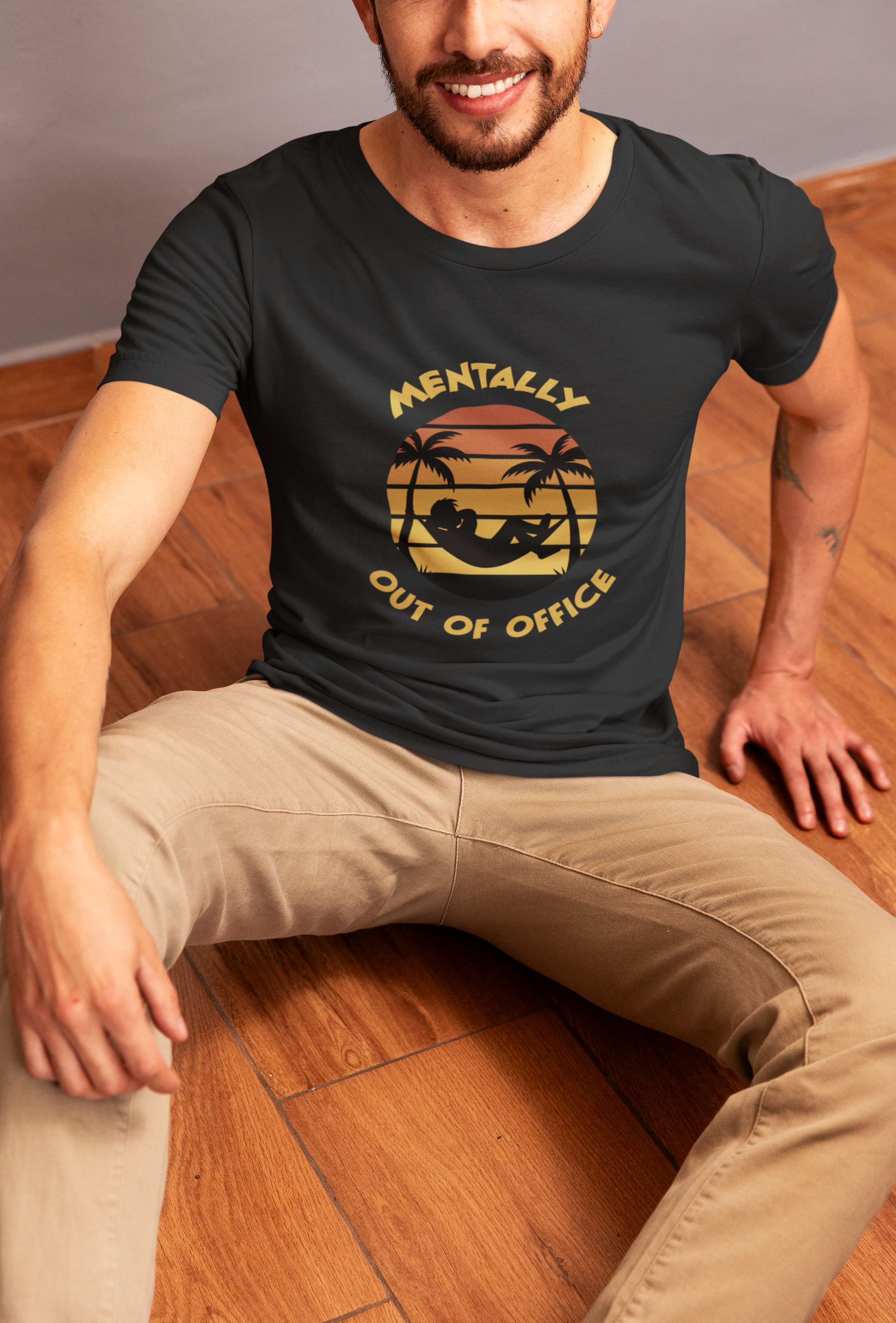 Mentally Out Of Office Tee Man wearing a black t-shirt with a graphic design and text, sitting on a wooden floor.