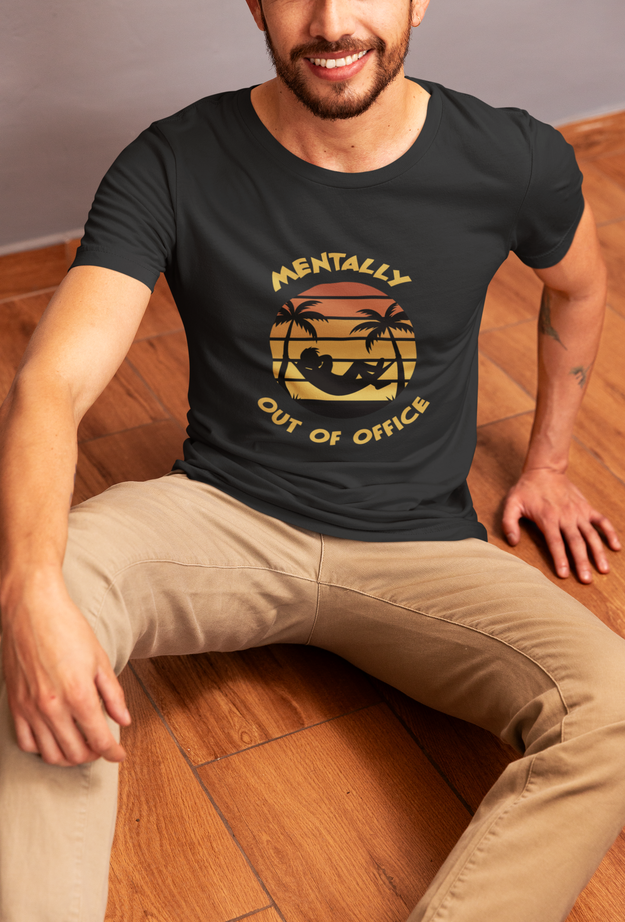 Mentally Out Of Office Tee Man wearing a black t-shirt with a graphic design and text, sitting on a wooden floor.