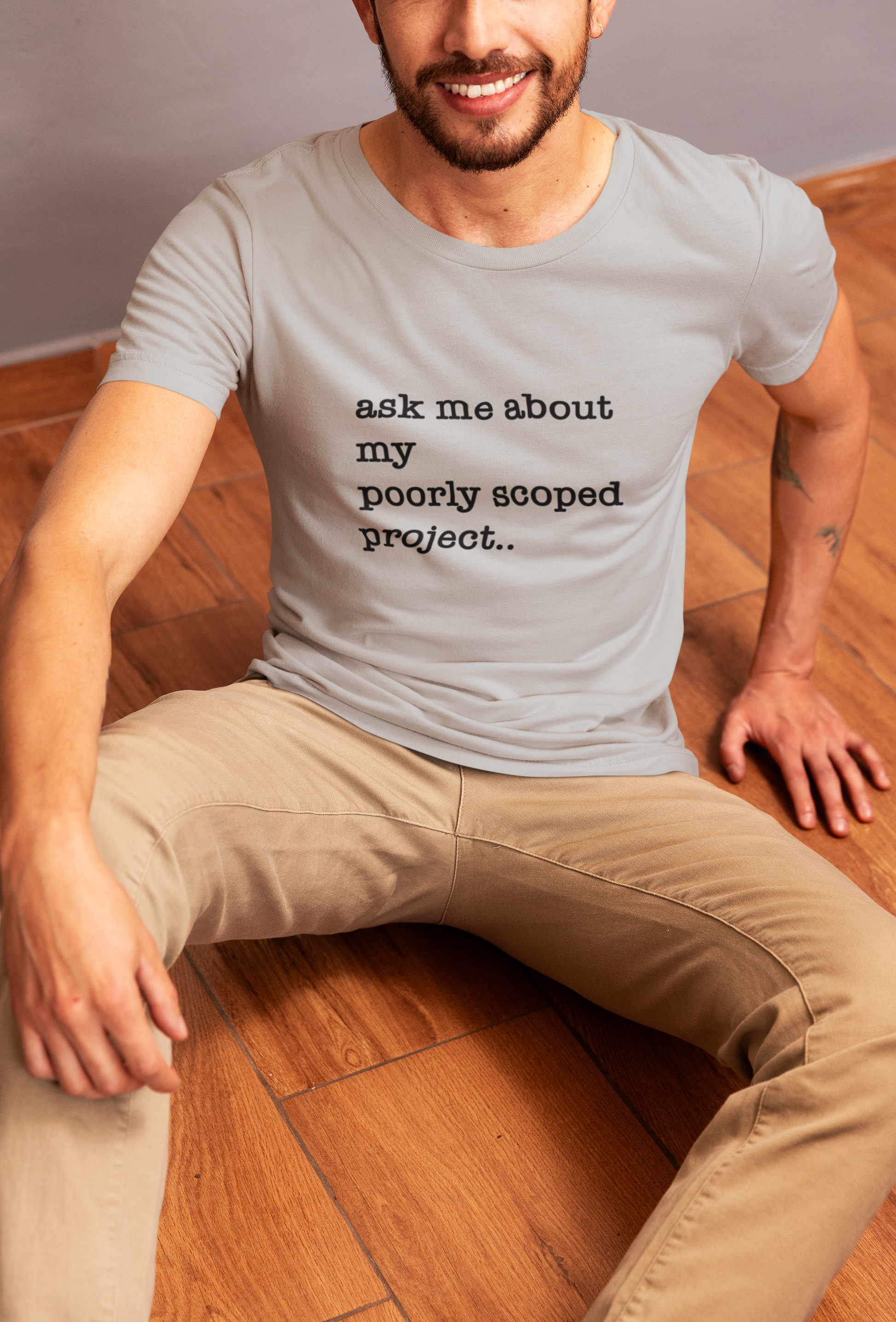 Organic Cotton Tee: Ask Me About My Poorly Scoped Project Man wearing a black t-shirt with a graphic design and text, sitting on a wooden floor.
