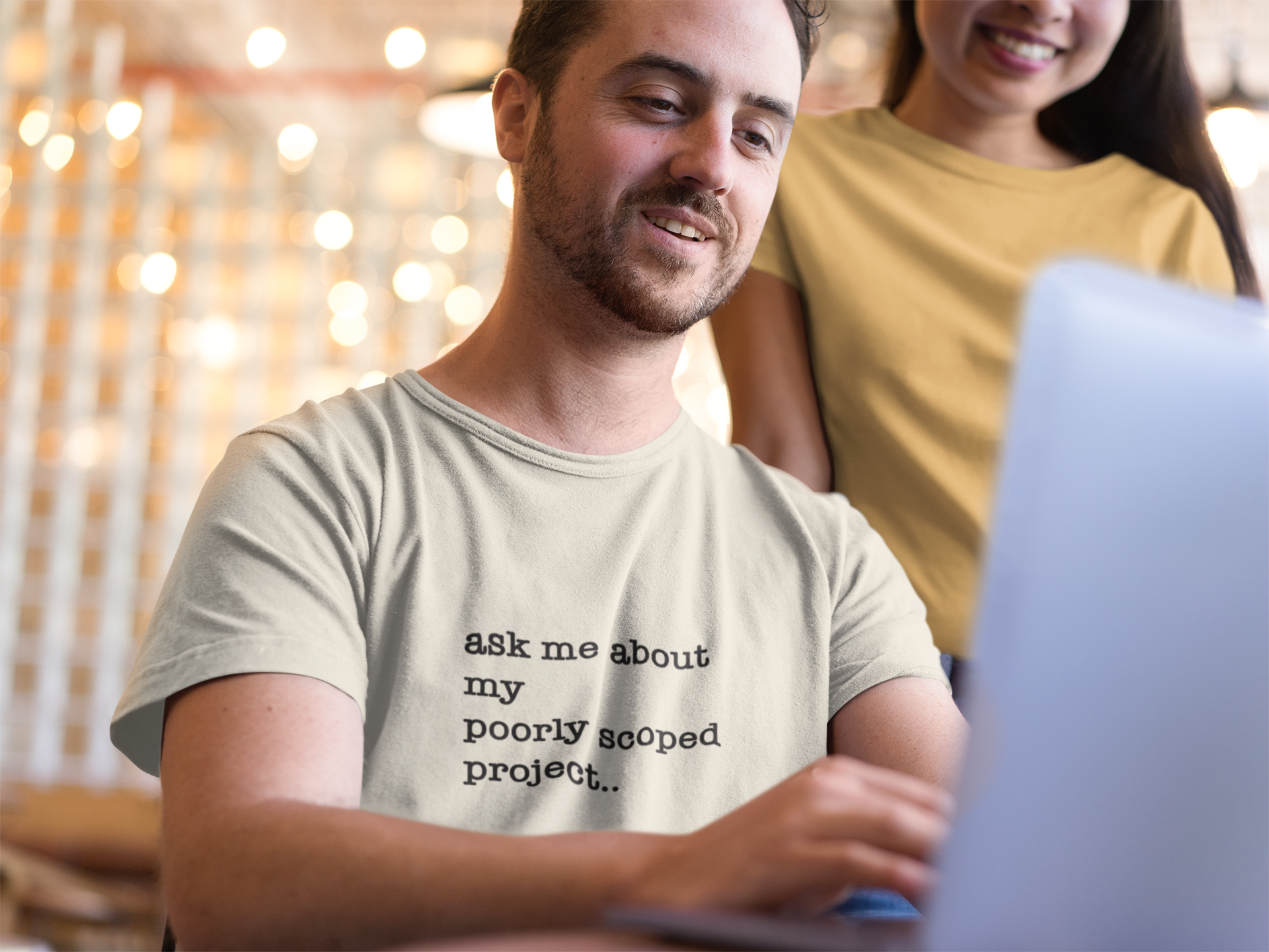 Organic Cotton Tee: Ask Me About My Poorly Scoped Project. Man wearing a shirt with text, sitting in front of a laptop with a woman standing behind him.