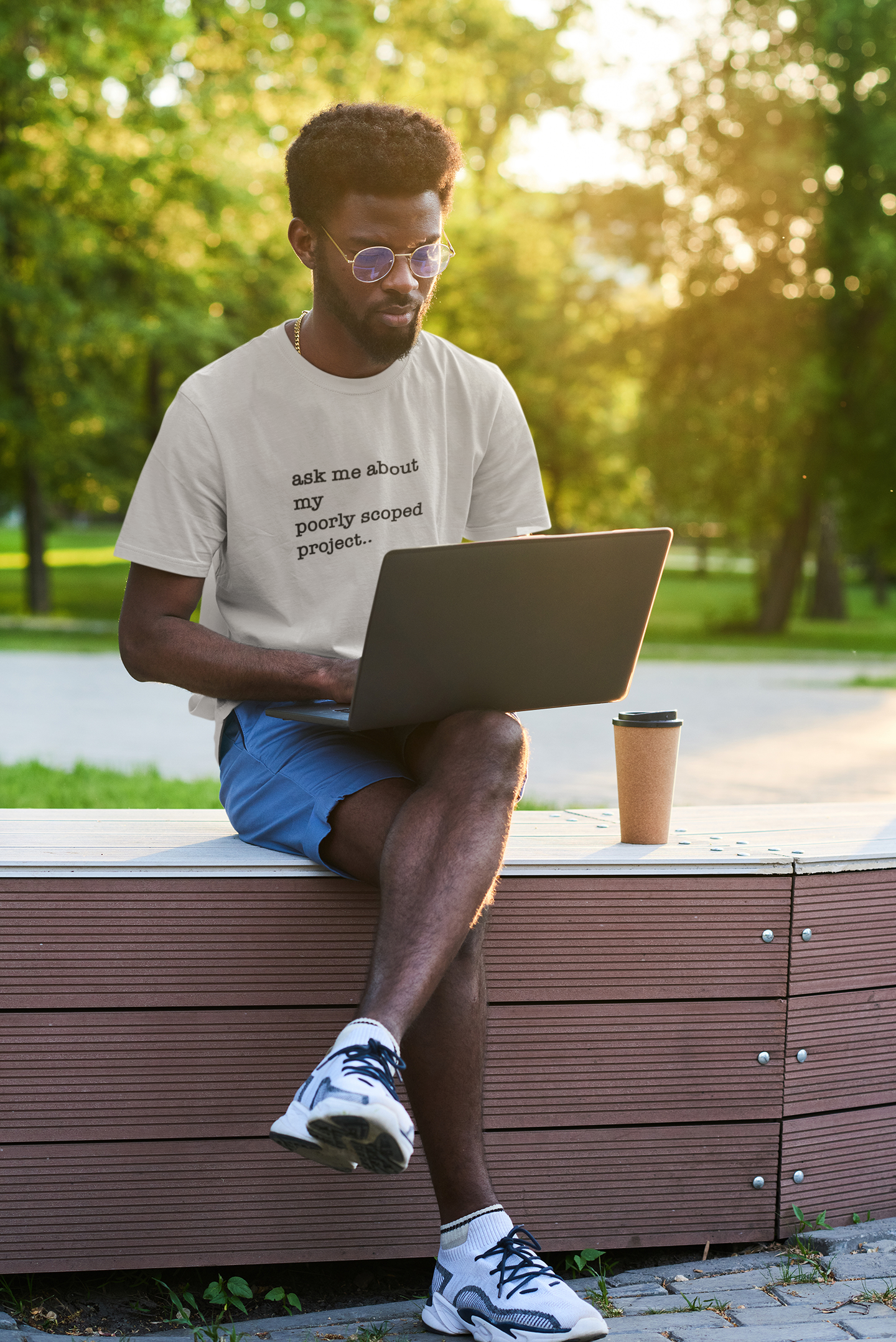 Organic Cotton Tee: Ask Me About My Poorly Scoped Project Man sitting on a bench outdoors using a laptop with a cup of coffee.