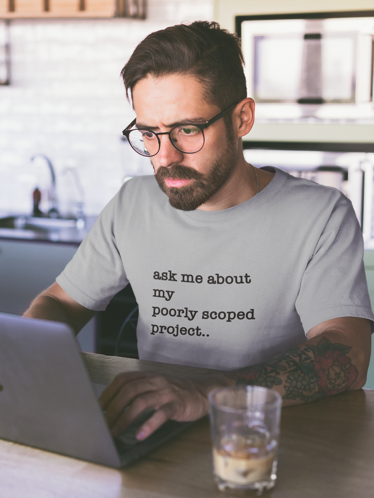Organic Cotton Tee: Ask Me About My Poorly Scoped Project Man wearing a shirt with text working on a laptop in a kitchen.