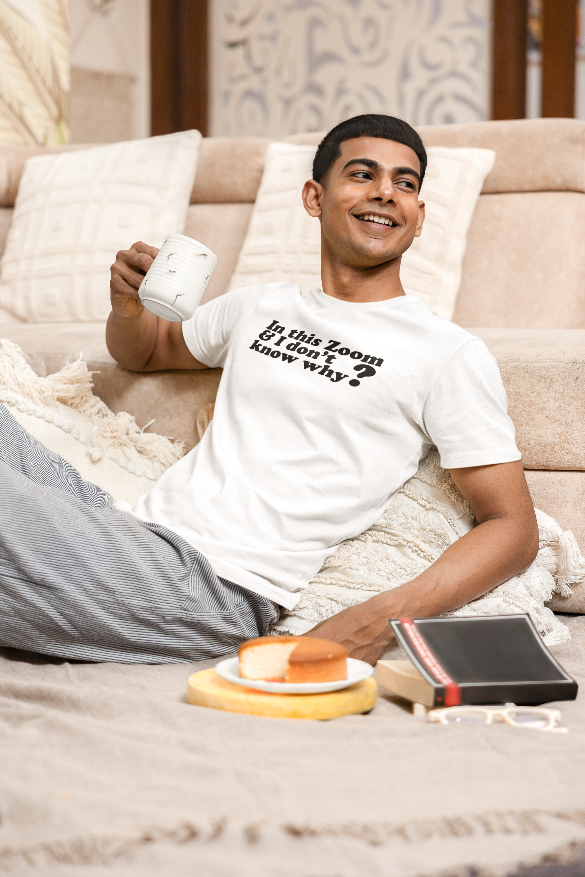 Organic Tee I'm In This Zoom Don't Know Why Man sitting on a couch holding a mug with a white shirt and gray pants, smiling.