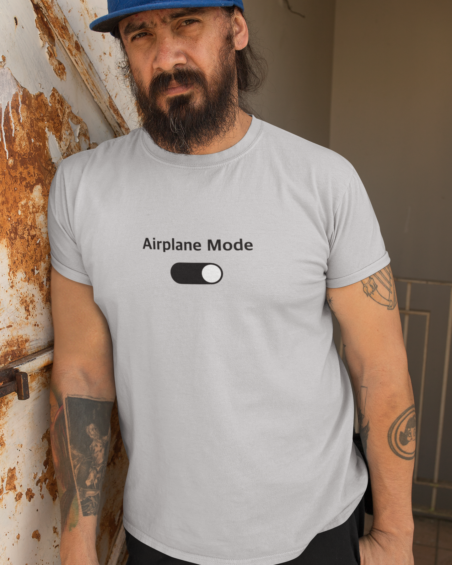 Man wearing a gray t-shirt with 'Airplane Mode' text and toggle graphic, leaning against a wall.
