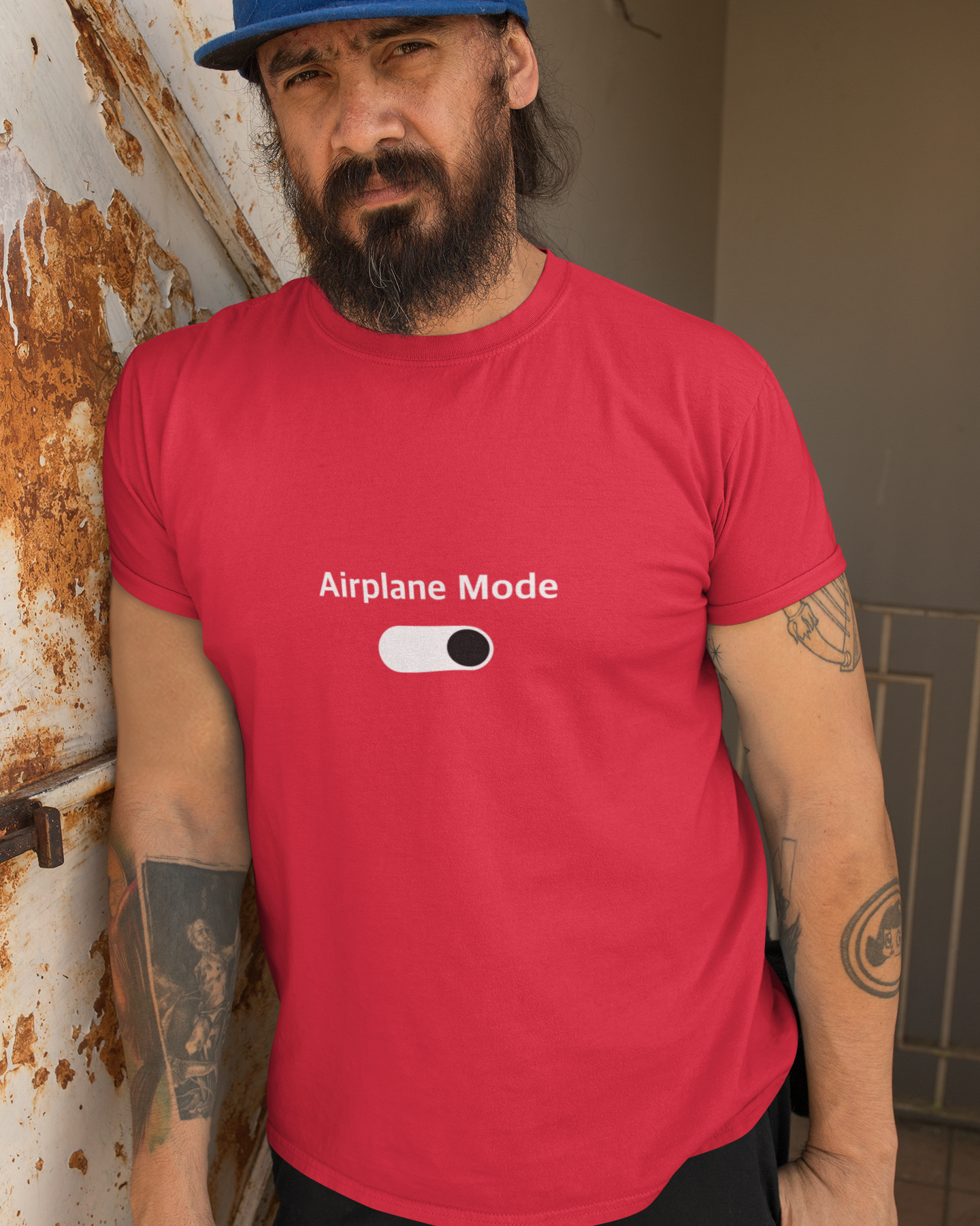 Man wearing a red t-shirt with 'Airplane Mode' graphic, standing against a rusty metal wall.