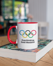 Mug with Olympic rings and 'Overthinking Gold Medalist' text on a stack of books.